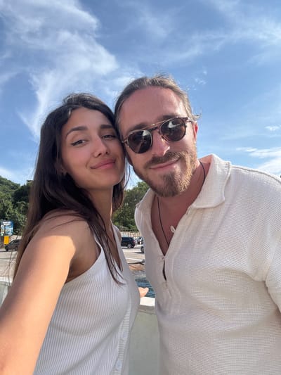 In this selfie, a man and a woman stand side-by-side against a backdrop of a bright blue sky with wispy white clouds. The woman, on the left, has long dark hair and is wearing a white ribbed tank top. She is looking towards the camera with a slight smile. The man, on the right, has medium-length brown hair, a beard, and is wearing sunglasses and a white collared shirt. He is also smiling at the camera. In the background, there are some trees and buildings, suggesting an outdoor urban or coastal setting. There are cars visible on a road behind them, and a body of water can be seen to the right. The overall impression is of a candid, happy moment captured outdoors. The location is identified as Prčanj, Montenegro.