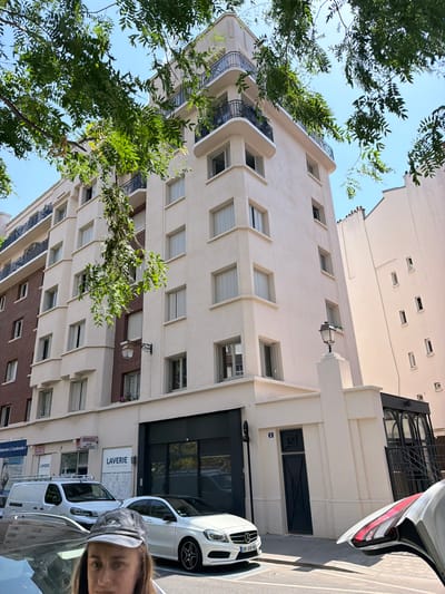 The image shows a street view in Courbevoie, France, with a modern apartment building in the background. The building is multi-story with white facade and balconies with dark railings. In front of the building, there is a row of shops, one of which is labeled "LAVERY" (laundry). A white van and a white Mercedes-Benz sedan are parked on the street. The foreground of the image is occupied by the upper body of a person wearing a dark baseball cap, partially obscuring their face. The person is looking towards the camera. The scene is illuminated by natural daylight, suggesting it was taken during the day. Lush green foliage from trees is visible in the upper part of the image, partially obscuring the building and the sky. The sky is clear and blue. The overall impression is a snapshot of urban life on a sunny day.