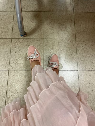 This is an eye-level, overhead shot of a person's legs and feet wearing pink canvas shoes and a flowing pink dress, standing on a tiled floor. A metal chair leg with a blue base is visible to the left. The scene appears to be indoors, possibly a public area given the tiled floor and chair. There is no indication of the location being specifically in Katunayake, Sri Lanka. The lighting suggests an indoor environment with artificial light.