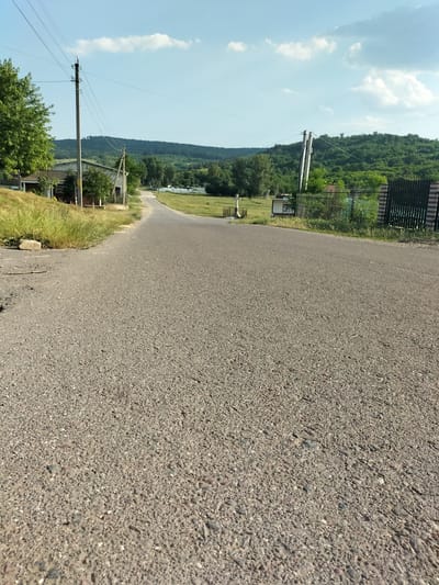 The image depicts a rural road winding through a landscape under a bright, partly cloudy sky. The road itself is paved with asphalt and occupies the foreground, with its texture of small stones and gravel prominently displayed. To the left of the road, a grassy embankment slopes upwards, leading to a cluster of buildings, including what appears to be a house with a light-colored brick facade and a darker roof. A tall, utility pole with wires stretches upwards beside the building, a common sight in rural areas. Beyond the buildings, a line of trees adds to the scenic view, with a lush green hillside in the distance forming the backdrop.
On the right side of the road, a black metal fence with brick posts lines the property. Further down the road, more trees and vegetation are visible, suggesting a well-treed environment. In the far distance, rolling hills covered in dense green foliage dominate the horizon, hinting at a mountainous or hilly terrain. The presence of utility poles and the architectural style of the buildings suggest a location in a rural village or settlement.
The time of day appears to be daytime, likely late morning or early afternoon, given the bright sunlight casting subtle shadows and the clear visibility. The weather seems pleasant, with a gentle breeze suggested by the scattered clouds in the blue sky. There are no people visible in the image, and no discernible text. The overall mood is peaceful and serene, characteristic of a quiet countryside setting.