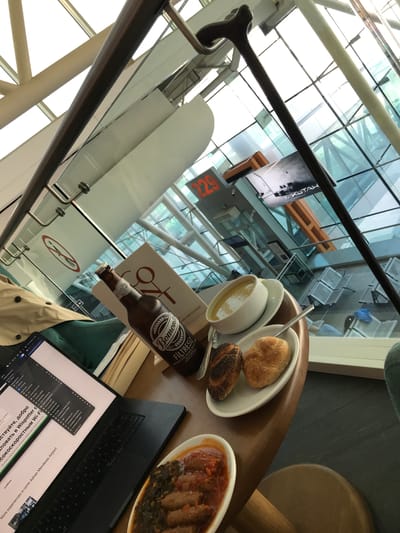 The image displays a table setup within a lounge or cafe area of Izmir Adnan Menderes Airport in Gaziemir, Turkey. The perspective is from behind a seated individual, looking out through large, multi-pane glass windows revealing the airport's interior architecture, including steel beams, distant seating arrangements, and a digital gate display showing "229." An advertisement or sign featuring "KUTAH" and "BERMİK" is visible beyond the window.

On a wooden table, a black laptop is open, displaying a webpage with text in Russian and English, specifically mentioning "Wispotter" and "Izmir Adnan Menderes Airport" Wi-Fi services. Beside the laptop, an open brown glass bottle of "Bomonti Filtresiz" beer is present. A white menu or document is partially obscured behind the beer bottle. A meal consisting of a white bowl of yellow soup with a spoon, a white plate holding two bread rolls (one seeded, one plain), and a white oval dish containing meat (possibly köfte) with green vegetables and red sauce is arranged on the table. A tan jacket or coat is draped over a chair to the left. A dark brown wooden cane with a curved handle leans against a metal railing structure in the upper right portion of the frame. A "no smoking" symbol is visible on a glass partition near the table.