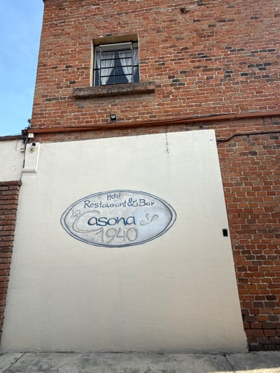 A low-angle, outdoor photograph depicts a building corner in Tepeji de Ocampo, Mexico. The corner comprises a section of brick wall with a barred window, adjacent to a white plastered wall. The wall is adorned with an oval-shaped sign in blue lettering that reads "Hotel Restaurant & Bar La Casona 1940." An electrical conduit runs down the brick wall near the corner. The sky is visible in the lower left corner.
