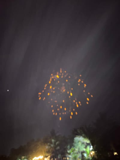 A nighttime scene in Habaraduwa, Sri Lanka, depicts a blurry display of what appears to be fireworks or sky lanterns against a dark, hazy sky. Numerous small, out-of-focus orange lights are scattered across the upper-middle portion of the frame, suggesting an event involving illumination in the sky. To the right, a blurred green mass with some bright white and yellow highlights indicates foliage, possibly trees, with some artificial lights, perhaps festive decorations, visible. The overall impression is one of a celebration or gathering taking place outdoors at night. The ambient light is dim, and the focus is soft, creating an atmospheric but indistinct view of the activity.