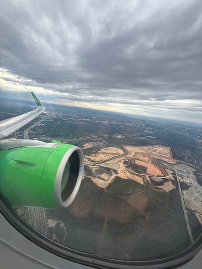 The image shows an aerial view from inside an airplane, likely during descent or ascent. Outside the window, there's a green engine of a Viva Aerobus plane and a wing with the "Viva" logo. The landscape below features a mix of urban and natural environments, including what appears to be an open pit mine, with red soil contrasting with the green vegetation. The urban area shows buildings and roads, including highways and clusters of buildings. The sky is covered with gray, overcast clouds, suggesting a cloudy day. The location is possibly near San Antonio, United States of America, based on the landscape's characteristics and presence of large-scale open pit mining.