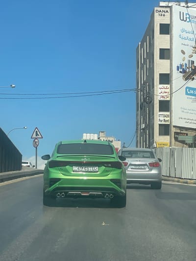 The scene depicts an urban street in Amman, Jordan, specifically identified near the DANA 126 building on Queen Rania Al Abdullah Street. The perspective is from within a vehicle looking forward.
In the foreground, a bright green Kia Forte sedan with Jordanian license plate 49-46102 is visible from the rear. It features a rear spoiler and dual exhaust tips. Directly behind this vehicle is a silver Hyundai Elantra sedan, also viewed from the rear, bearing Jordanian license plate 18-65276. Both cars are on an asphalt road.
To the left of the road, a grey concrete barrier runs alongside, and two road signs are visible: a triangular 'merge from right' sign and a circular 'no U-turn' sign. Overhead power lines traverse the blue, clear sky.
On the right side of the image, a light-colored, multi-story building stands, labeled "DANA 126" at its upper section. Attached to this building is a vertical banner displaying "مكاتب ومعارض للايجار" (Offices and showrooms for rent), a phone number (0777304288), and "For Rent" in English. Adjacent to this, a large advertising billboard for "Windsor Brokers" is partially visible, with Arabic text. A corrugated metal construction barrier is present below the billboard. No individuals are visible in the image.