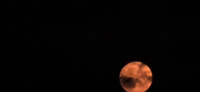 The image shows a full, orange-hued moon against a dark, starless sky. The moon's surface displays visible craters and cloud-like formations, suggesting it is either partially obscured by atmospheric haze or the photograph has been processed to enhance these features. The lighting is dim, and the overall scene is a close-up of the celestial body. There is no indication of any specific location in Mazyr, Belarus, nor are there any people, objects, or actions present in the image.