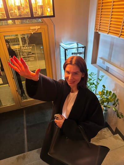 A woman wearing a black coat and white shirt smiles while holding a phone in her left hand. Her right hand is extended forward with red nail polish visible. The woman is standing in front of a glass door with a restaurant sign. A black tote bag rests on her shoulder. A green plant is visible in the background, along with indoor lighting fixtures. The scene appears to be outside the restaurant.
