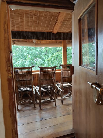 The image shows an open wooden door leading to a covered outdoor seating area in Prini, India. The seating area has a wooden floor and a slatted bamboo ceiling. Three woven rattan chairs are positioned on the floor facing a window that looks out onto a lush green trees. A brass door latch is visible on the exterior of the wooden door.