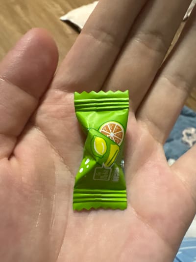 An extreme close-up shot shows a person's hand holding a green, individually wrapped candy. The candy wrapper has a serrated edge at the top and bottom and features an illustration of a sliced orange and a lime. Text in an East Asian script is visible on the lower portion of the wrapper. The hand is open, with the candy resting in the palm, and the fingers are curled slightly. The background is blurred, showing a textured surface and a hint of fabric with a pattern. The lighting appears to be artificial and overhead, casting subtle shadows on the hand.