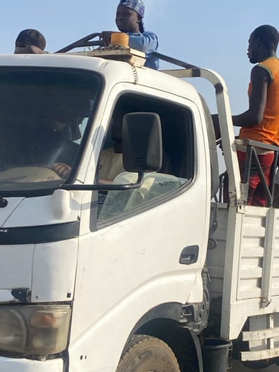 This image depicts a white medium-duty truck operating on a bright, clear day in Katcha, Nigeria. Multiple individuals are present on and within the vehicle. A male subject, wearing a blue long-sleeved shirt and a patterned cap, is positioned on the cab's roof, leaning slightly forward with hands on a custom metal rack. An orange cylindrical object is affixed to this rack. To the left of this subject, a dark head covering indicates the presence of another individual on the roof. Through the driver's side window, a person wearing a light-colored shirt is partially visible inside the cab. Standing in the white metal-railed bed of the truck, another male subject is visible, wearing an orange sleeveless shirt and dark shorts, facing the front of the vehicle. The truck's front section, including a headlight and a tire with "AUSTRO" branding, is partially visible. The background consists solely of a clear sky, indicating outdoor conditions.