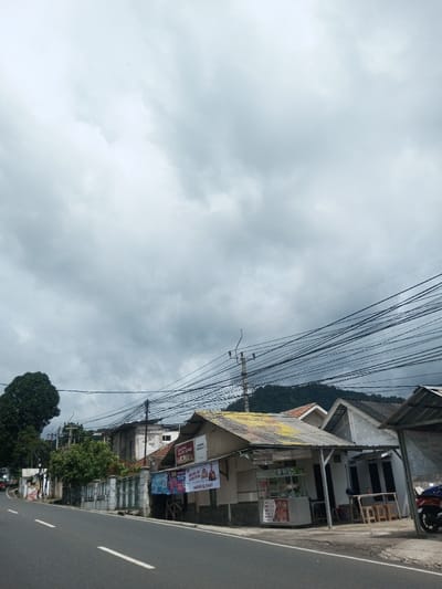 A street view beneath a cloudy sky features buildings, wires, and vegetation. A road runs alongside the buildings, one of which appears to be a small shop with advertisements. A mountain ridge is visible in the distance, partially obscured by the wires. A tree stands to the left of the buildings. The scene suggests a rural or suburban environment.