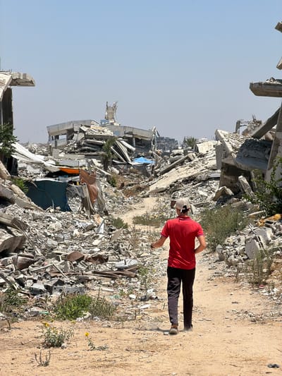 The image depicts a desolate scene in Jabalia, Palestinian Territories, characterized by widespread destruction. In the foreground, a person wearing a red shirt, black pants, and a light-colored cap walks on a dirt path. The path is bordered on the left by a vast expanse of rubble, consisting of shattered concrete, rebar, and debris from what appear to be collapsed buildings. The sheer scale of the destruction is emphasized by the towering piles of concrete slabs and twisted metal.
In the background, more of the devastated urban landscape is visible, with remnants of damaged structures stretching into the distance under a clear, bright sky, suggesting it is daytime and the weather is fair. The presence of scattered vegetation, including small green plants and wildflowers, growing amidst the ruins offers a subtle contrast to the surrounding destruction. Tents and tarps are visible within some of the collapsed structures, possibly indicating makeshift shelters. The overall atmosphere conveyed is one of devastation and the aftermath of conflict. No visible text is present in the image.