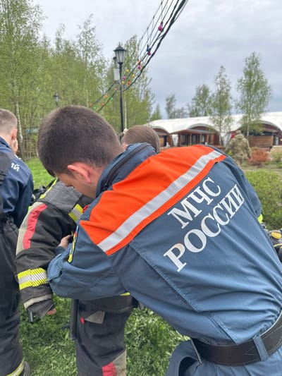 In Grebnevo, Russia, a first responder wearing a dark blue uniform with orange stripes and the Russian acronym for Ministry of Emergency Situations (МЧС РОССИИ) is bending over a person in a firefighter's turnout coat. The two are located on a grassy area near a lamppost with strings of lights. A building with white curved supports is visible in the background.