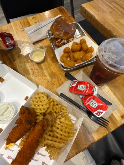 The image shows a wooden table with various food items and a drink, suggesting a meal. On the table, there is a burger on a metal tray with a side of tater tots. Next to it, a plastic cup contains a dark beverage with foam. In the foreground, a white takeout box holds waffle fries and what appear to be chicken tenders. There are also several small containers of dipping sauce and packets of ketchup. Utensils, including a fork and knife, are also visible. The restaurant's branding, "Bruxie," is visible on the takeout box. The overall scene suggests a casual dining experience. The provided context indicates the location is Santa Monica, United States of America.