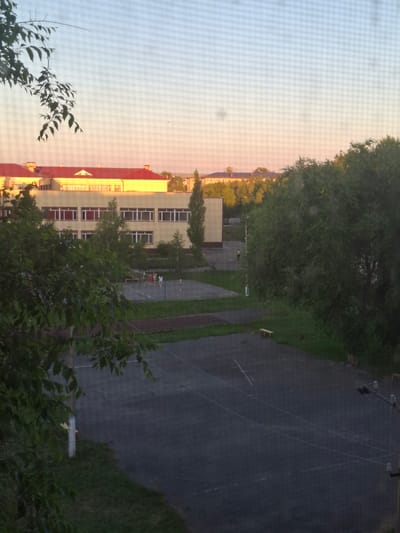 The image depicts an outdoor scene, likely a schoolyard or public space, viewed from a window with a mesh screen. The perspective is from a high vantage point, looking down and across. In the foreground, lush green foliage, possibly trees and bushes, partially obscures the view.
The midground features a multi-story building with a red roof and cream-colored walls, punctuated by numerous windows. Sunlight casts a warm glow on the building, suggesting it is either early morning or late afternoon. Adjacent to the building is a dark asphalt area, resembling a sports court or playground, with white lines marking boundaries. A few scattered objects, possibly benches or equipment, are visible on this surface. Further in the background, more greenery and distant structures can be seen.
Several individuals, appearing to be children, are gathered on the court, their activity unclear due to the distance and resolution. The sky above is a pale, gradient hue, transitioning from a bright, light color to a warmer, pinkish tone near the horizon, reinforcing the impression of dawn or dusk. The overall atmosphere suggests a calm, ordinary day. No text is visible in the image. The location is Jitiqara, Kazakhstan.