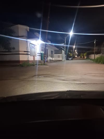 The image captures a street scene at night in Yeghegnadzor, Armenia, illuminated by streetlights and light emanating from buildings. In the foreground, the hood of a vehicle is visible, suggesting the image was taken from inside a car. The street is paved with asphalt and shows some wear and tear, with visible tire marks and scattered debris.
To the left, a building with a white facade and dark horizontal accents is present. It features large windows, some of which are illuminated from within. Adjacent to this building, there is a patch of green vegetation. Further down the street, another building, seemingly residential or commercial, can be seen.
The street itself is lined with trees and other vegetation on the right side. Several streetlights are visible, casting bright beams of light that create streaks and lens flares in the image due to the night conditions. A dark-colored car is parked on the right side of the road in the middle distance. The overall environment suggests a quiet urban or suburban street at night.
