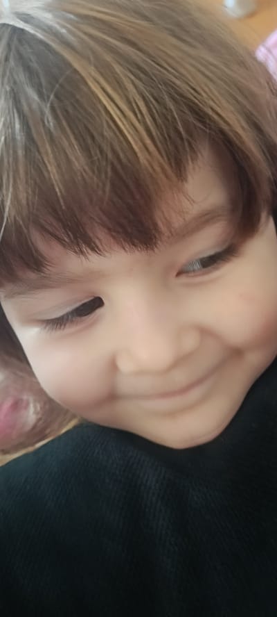 A close-up, head-and-shoulders shot shows a young child with light brown hair and fair skin. The child's face is turned slightly to the right, and they are looking down and to the right with a subtle smile. The background is blurred, showing hints of pink on the left and a dark, textured fabric (likely clothing) in the foreground and on the right. The image appears to be taken indoors, with no discernible elements of the city of Хохлы, Russia, visible.