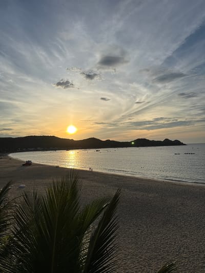 The image depicts a scenic sunset over a beach in Song Cau, Vietnam. The sun, a bright orb, is positioned in the center of the frame, casting a golden glow across the sky and water. Wispy clouds, tinged with orange and pink hues, stretch across the upper portion of the image. A gentle wave breaks on the sandy shore in the foreground.
In the mid-ground, a few scattered boats dot the calm sea, their silhouettes softened by the fading light. The coastline curves gently, bordered by a range of dark, tree-covered hills. Along the beach, a red vehicle with a white canopy is parked, and two individuals are walking near the water's edge.
The foreground is partially obscured by the dark silhouette of palm fronds, adding a tropical feel to the scene. The overall impression is one of tranquility and natural beauty, characteristic of a coastal sunset in this region.