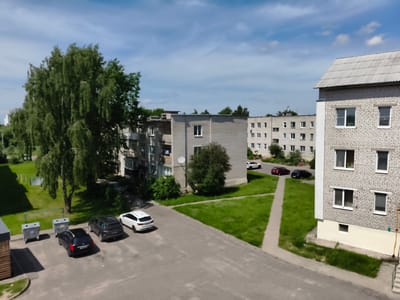 The image depicts an exterior view of a residential area in Myadzyel, Belarus. Multiple apartment buildings constructed with light-colored bricks are visible, along with a few parked cars on an asphalt lot and a paved walkway. Lush green trees and grass are interspersed throughout the scene, and the sky is partly cloudy with blue patches. In the foreground, to the left, are several industrial-style garbage bins. The overall impression is that of a typical urban or suburban setting on a clear day.