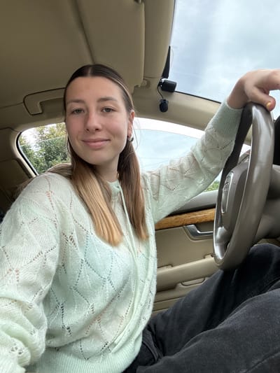 A young woman with blonde hair and light blue eyes is sitting in the driver's seat of a car, looking at the camera. She is wearing a light mint green crocheted sweater over a light blue bra. Her left arm is resting on the steering wheel, which has the Audi logo on it. She has on dark wash jeans and is wearing hoop earrings. The car's interior is visible, with tan leather seats and wood trim. Outside the car, trees and a cloudy sky can be seen through the windows. There is a small black microphone clipped to the car's headliner near the windshield. The car appears to be parked, as there is no indication of movement or driving. The image was taken in the city of Yartsevo, Russia.