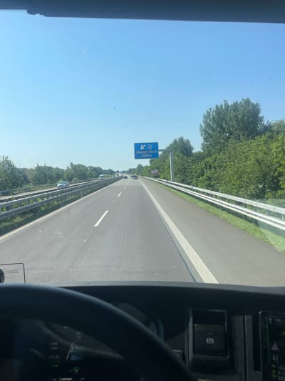 This image, taken from the perspective of a vehicle's cabin, captures a view of a highway in Haren (Ems), Germany. The scene is dominated by a clear blue sky and lush green trees lining the sides of the road. A white guardrail separates the highway from the trees.
The highway itself is asphalt, with white lane markings visible. Several cars are driving on the road, suggesting moderate traffic. A blue highway sign is prominently displayed, indicating "Meppen-Nord" and a distance of "2000 m" (2 kilometers). This sign also features the number "21" within a blue circle, likely representing the highway number.
In the foreground, part of the vehicle's interior is visible, including a steering wheel and a dashboard with various indicators. A button with a "P" symbol, possibly indicating a parking brake, is also present. Digital displays on the dashboard show information like speed ("120 km/h"), time ("13:00"), and other numerical data. The bright sunlight suggests it is daytime, likely midday. The overall impression is one of a normal, uneventful drive on a highway under clear weather conditions.
