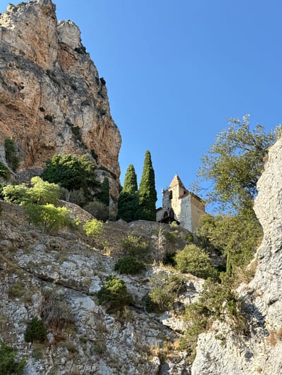 The image shows a steep, rocky cliff face with a small, stone church perched precariously on a ledge near the top. The church has a bell tower and a pitched roof, and appears to be made of rough-hewn stone blocks. Cypress trees and other greenery are scattered amongst the rocks, with a few trees growing in front of the church. The sky is a clear, bright blue. The terrain is rugged and uneven, with many loose rocks and sparse vegetation. The location is identified as Moustiers-Sainte-Marie, France. The scene conveys a sense of isolation and permanence. There are no people visible in the image.
