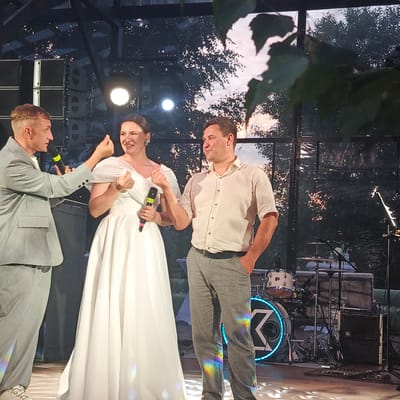 In the city of Svetloe, Russia, a man in a light grey suit, holding a microphone, gestures towards a woman in a white wedding dress. The woman also holds a microphone. Beside her, a man in a beige shirt and grey pants stands with his hand in his pocket, looking to the side. They are on a stage with a drum set and stage equipment in the background, set against a backdrop of trees and a sunset visible through glass walls. Rainbow-colored light reflections are visible on the floor and the subjects' clothing.