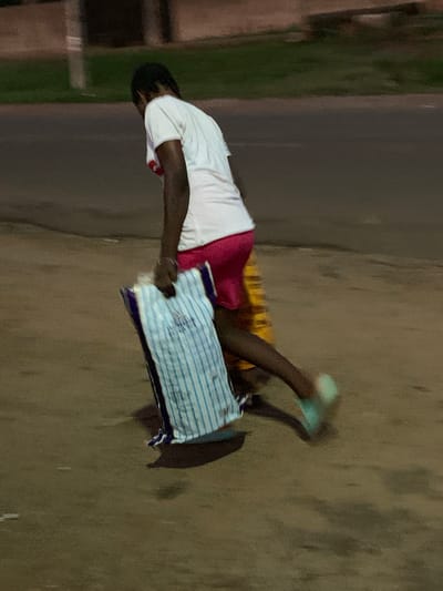 A person wearing a white shirt and pink shorts is lying on a striped pillow on a concrete surface. The person is wearing what appears to be light green footwear. The location appears to be outdoors, possibly in an urban area, but the specific location in Gbeleko, Nigeria, cannot be determined from the image alone. The environment includes a dark asphalt surface and some blurred greenery in the background. The person's actions are limited to lying on the pillow. There are no apparent interactions with other people or objects.