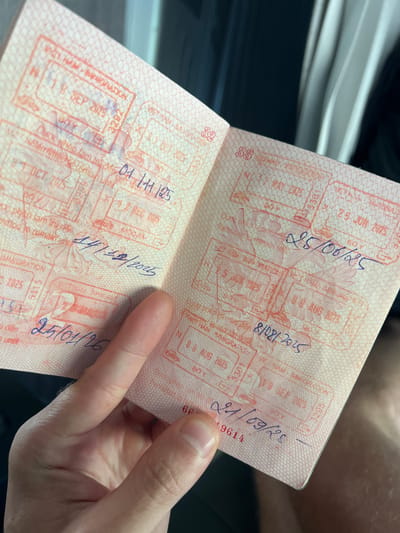 A person is holding open a passport, displaying several visa stamps. The passport pages are printed with a fine, pink, patterned background. The stamps are primarily red ink and feature an ornate design, with specific dates and abbreviations over them written in blue ink. The dates visible on the stamps are: 01.11.125, 14/12/2025, 25/01/26, 25 JUN 2025, 25/06/25, 8/08/2025, and 21/09/23. The stamps include some information related to immigration. Text in the background is "Duoc Nico lam to". The stamps feature a location within Vietnam; Kon Ra Kiah is not explicitly stated in the stamp details.