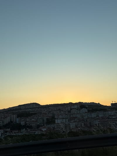 This is a vertical, eye-level, full shot photograph of the city of Loures, Portugal, taken at dusk. The sky is a gradient of clear blue at the top, transitioning to a soft yellow near the horizon where the sun has set. The city is built on a sloping hill, densely packed with buildings of various sizes and styles, predominantly apartment buildings with tiled roofs. Some taller, modern buildings are visible on the crest of the hill, along with communication towers. The foreground features a dark, blurred railing and some indistinct foliage, suggesting the photo was taken from an elevated vantage point, possibly a road or bridge. There are no people visible in the image. The overall scene depicts a sprawling urban landscape under a serene evening sky.
