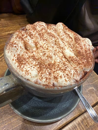 A close-up, angled shot shows a mug of hot chocolate topped with whipped cream and dusted with cocoa powder. The mug is a light blue-gray ceramic and sits on a matching saucer on a wooden table. A stainless steel spoon rests partially on the saucer and mug. A person's shoulder and arm are visible in the upper left, suggesting the beverage is being enjoyed at a table. The location within Mole Valley, United Kingdom cannot be determined from this image.