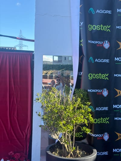 The image depicts an outdoor scene with a red curtain on the left and a large black banner on the right. The banner features the logos and text "AGGRE", "gostex", and "MONOPOLY WIN". A potted plant with green leaves is positioned in front of the banner and to the right of the curtain. In the background, a mirror reflects a young couple taking a selfie. The sky is blue with a few clouds, and a power line tower is visible in the distance. The scene suggests an event or promotional setup.