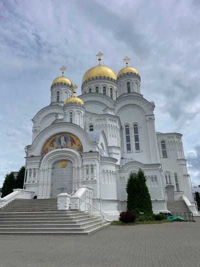 A large, white Orthodox cathedral with golden domes stands under a cloudy sky. The cathedral has multiple domes topped with crosses. The facade features intricate architectural details, including arched windows and decorative moldings. A mosaic depicting a religious scene is situated above the main entrance. A wide set of stone stairs leads up to the entrance, with white railings on either side. Green trees and landscaping are visible in front of the cathedral. The ground in front of the stairs is paved with gray bricks. The scene appears to be a still, exterior view of a religious site. The location is the Seraphim-Diveyevo Monastery in Diveyevo, Russia.