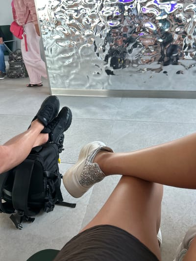 This media file features a close-up, overhead view of two individuals' legs and feet, with a focus on their footwear and a backpack. One person is wearing black athletic shoes and is positioned with their legs crossed, resting on a black backpack. The other person, partially visible, has on a sparkly white slip-on shoe and their legs are also crossed, one foot resting on the other's ankle. The setting appears to be indoors, possibly an airport or a waiting area, indicated by the tiled floor and a large, reflective, textured wall in the background. The lighting suggests daytime. No text is visible in the image. The scene evokes a sense of casual waiting or resting.