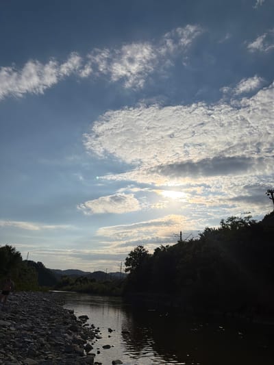 The image depicts a wide river flowing through a landscape of rolling hills covered in dense, dark green foliage. The sky above is a mix of blue and white, with scattered clouds, some of which are illuminated by the setting or rising sun, creating a bright glow in the center of the frame. Power lines and utility poles are visible on the right side of the riverbank, against the backdrop of the trees and hills. On the left bank, a person, appearing to be a male wearing dark shorts, stands on a rocky shore. The river reflects the sky and surrounding scenery, with ripples on its surface. The overall scene conveys a sense of natural beauty and a tranquil, possibly rural, environment. The location is identified as the city of Krasnoye, Russia.