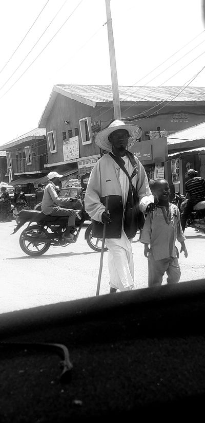 In this black and white photograph, several individuals are gathered on a street in Kinkinau, Nigeria. The scene is likely outdoors, given the presence of a building with windows and the open sky with overhead wires. The primary focus is on a person wearing a wide-brimmed straw hat, a light-colored jacket, and white trousers. This individual is holding a stick and appears to be interacting with a cart or a motorcycle.
Another person, wearing a patterned shirt, is seated and looking upwards with a broad smile, suggesting a moment of amusement or conversation. Behind them, another individual in traditional clothing is visible. The presence of a motorcycle with the word "HLX" visible on it, along with a "Smartcash" advertisement on the building, suggests a contemporary, urban environment. The lighting indicates it's daytime, and the shadows suggest the sun is present.
Visible text includes "BLOCK A", and what appears to be "MOUNT ZION COMPUTER ACADEMY" on a sign. The overall atmosphere seems lively and social, with people engaged in daily activities on a bustling street. The composition is somewhat dynamic due to the tilted angle of the photograph, adding a sense of movement to the scene.