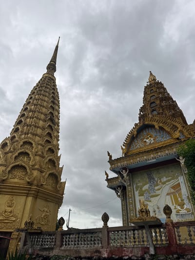 This image shows two ornate Buddhist structures under a cloudy sky. The structure on the left is a tall, golden stupa adorned with intricate carvings and multiple tiers. The structure on the right is a building with a decorative golden roof featuring a mural depicting figures and a scene that appears to be a procession or a ceremonial gathering. The foreground shows a weathered, decorative railing. The image is taken from a low angle, looking up at the structures. The location is identified as the city of Sneung, Cambodia.