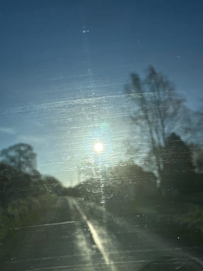 The image shows a view through a dirty window with streaks and smudges. The sun is visible through the window, appearing as a bright, circular light source. The reflection of trees and vegetation is also visible through the window. Due to the lack of specific information within the image, it is impossible to provide the exact location in Ruyton-XI-Towns, United Kingdom, nor is it possible to determine the presence of any people, interactions, or actions within the scene.