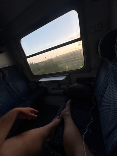 An interior shot of a train carriage shows a person's bare legs and hand resting on seats. The person is facing away from the camera, looking out of a window at a landscape. The sun is low in the sky, casting a warm glow over the scene. The seats are upholstered in a blue, patterned fabric. A black bag is visible on the seat in front of the person. The train appears to be traveling through a rural area. The location of Kameniak, Bulgaria is irrelevant to the image.