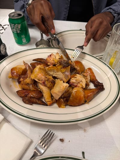 A photograph shows a white plate with a green rim containing what appears to be roasted chicken, partially carved and arranged. A person with dark skin is using a fork and knife to cut the chicken. A partially obscured green can of "Sparkling Water" is visible near the lower left edge of the plate. Utensils are scattered around the plate, and a white tablecloth is visible in the background. The photo was likely taken indoors. The image does not provide enough information to reliably determine if the location is in Qinhuai District, China.
