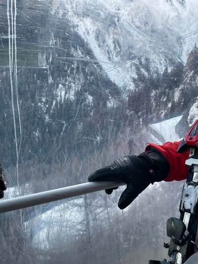 The image presents a close-up, first-person perspective from within what appears to be a ski lift cabin, looking out through a heavily scratched, transparent window. In the foreground, a person's left hand, covered by a black winter glove with a red cuff, is seen gripping a white or light grey ski pole. The brand name "REUSCH" is visible on the glove. Above the hand, part of a ski binding, colored red and metallic, is visible, suggesting the presence of a ski.

The background, viewed through the scuffed window, reveals a snowy, forested mountain landscape. Snow-covered evergreen trees are discernible, indicating winter conditions. The numerous scratches on the transparent surface create a textured, somewhat distorted view of the outside world. The lighting suggests it is daytime. The scene depicts an individual engaged in or traversing a ski area, consistent with a location like Courmayeur, Italy. The activity taking place is travel on a ski lift during a snowy winter day.