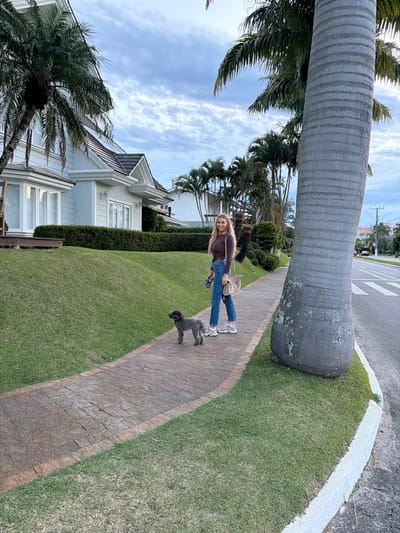 A woman with blonde hair walks a small grey dog on a leash along a paved sidewalk. The woman is wearing a brown long-sleeved shirt, blue jeans, and white sneakers. She has a tan shoulder bag. The dog is on a leash that the woman holds in her left hand. The sidewalk is bordered by a well-maintained green lawn and a paved street. In the background, there are houses and palm trees. The sky is cloudy. The location is identified as Florianópolis, Brazil.