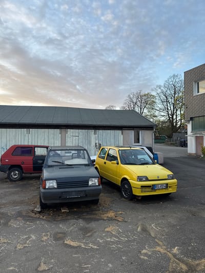 The image shows three cars parked on an asphalt surface beside a corrugated metal building, likely in a residential or commercial area. The cars are a yellow Fiat Cinquecento with the license plate "RS RD 4" facing right, a dark grey Opel Corsa facing right, and a red Opel Trio facing right. The yellow car has a German license plate, suggesting the location is in Germany. Based on the license plate starting with "RS," which indicates Remscheid, the cars are probably parked somewhere in the city of Remscheid, Germany. The corrugated building has a window visible. Beyond the building, trees are visible against a cloudy sky. The asphalt surface shows signs of wear and dirt. The image is rotated 90 degrees clockwise. There are no people visible in the image.