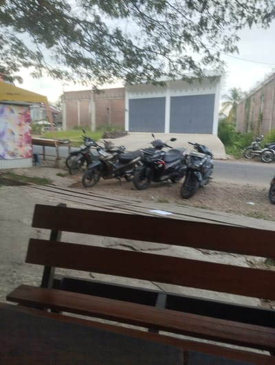 The image depicts a street scene in Mee, Indonesia, with several motorcycles parked on the side of the road. In the foreground, a wooden bench is visible, suggesting a seating area. Behind the bench, the motorcycles are lined up, with some appearing to be in use while others are stationary. The background shows a building with a large grey door and a brick wall. Trees with lush green foliage are present, contributing to the natural environment of the area. The sky is overcast, indicating a cloudy day. The overall impression is of a quiet, everyday setting in an Indonesian city.
