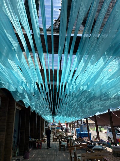 The image captures an outdoor covered walkway or street, viewed lengthwise, creating a strong sense of depth. A prominent feature is the overhead canopy, constructed from numerous long, translucent light blue fabric strips arranged in flowing, wave-like patterns. Bright sunlight filters through these strips, casting intricate patterned shadows on the ground and illuminating the fabric, highlighting its texture and movement.

The walkway below is lined on both sides with wooden structures, which appear to be market stalls, tables, and benches. On the right side of the path, a lone individual stands, dressed in dark clothing (black jacket/shirt, dark pants), facing away from the camera and looking further down the walkway. In the background, beyond the wooden structures, a street is visible, with a white car parked on the left. Various items, possibly goods for sale, are dimly visible on the stalls, and a red plastic stool along with a purple bucket are on the ground near the person.

The scene is set during a bright, sunny daytime, evidenced by the strong light and distinct shadows. The clear weather suggests good conditions for an outdoor market or leisurely stroll. This location, specifically Jiejinkou, China, appears to be a vibrant commercial or pedestrian area. No legible text is visible within the image, and the overall mood seems calm and observational.