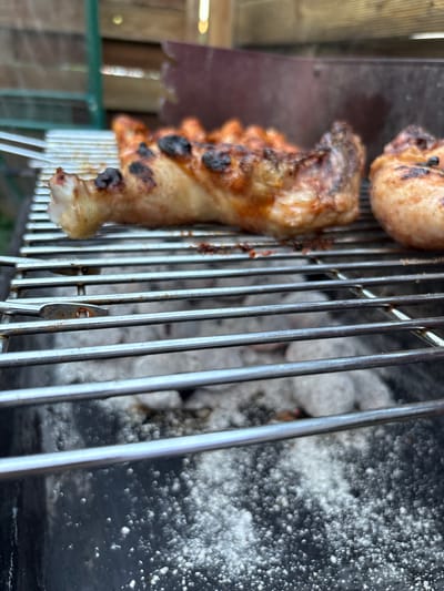 The image shows a close-up view of chicken pieces being grilled on a charcoal barbecue. The chicken is seasoned and has grill marks, with some parts appearing slightly charred. The grill grate is made of metal bars, and below it, glowing charcoal briquettes are visible. In the background, a wooden fence and some greenery can be seen, suggesting an outdoor setting. The image is taken from a low angle, looking up at the grilling food. There is no indication of the city of Delbrück, Germany in the image.