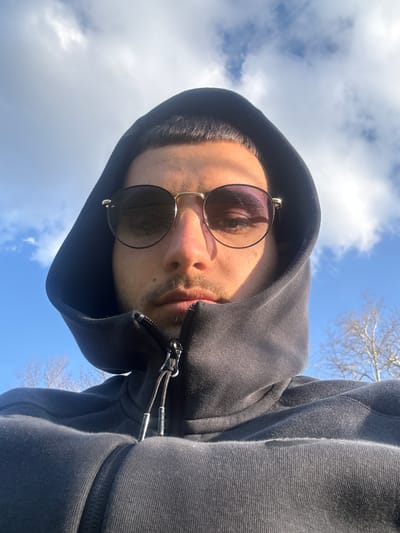 A man wearing sunglasses and a black hoodie is positioned in the center of the frame, looking down at the camera. The sky is visible in the background, featuring blue patches and white clouds. Bare branches are present in the upper right area. The photograph does not have sufficient information to determine its location in the city of Dushevo, Bulgaria.