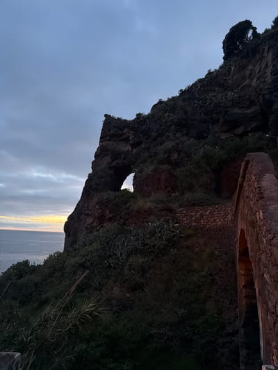 The image shows a rock formation with a hole in it, set against a cloudy sky. The location is Ponta do Sol, Portugal, based on the landscape. A stone structure is built along the top edge of the cliff. The rock formation is covered in green vegetation on its edges. The ocean is visible at the bottom of the image.