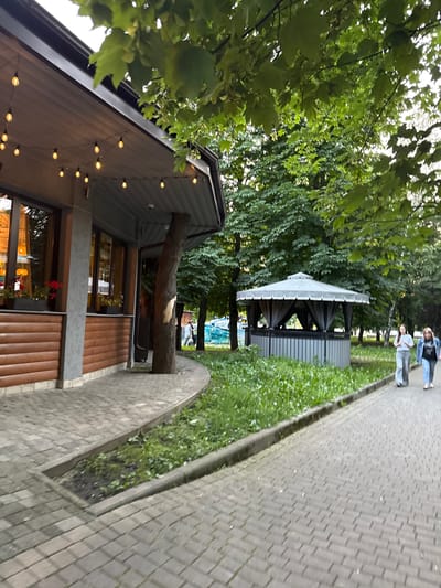 The image depicts an outdoor scene with a modern building featuring a sloped roof adorned with string lights. A tree trunk is visibly integrated into the structure, growing through an opening in the building. Adjacent to the building is a paved walkway, bordered by a strip of grass and trees. Further along the walkway, a gazebo with a white roof and dark curtains is visible, surrounded by lush greenery. In the background, a few individuals are walking along the path, and hints of other structures and trees suggest a park or outdoor recreational area. The lighting indicates it might be late afternoon or early evening, with soft natural light. The overall impression is a serene, possibly urban park setting with a touch of rustic charm from the tree and building design. No visible text is present in the image.