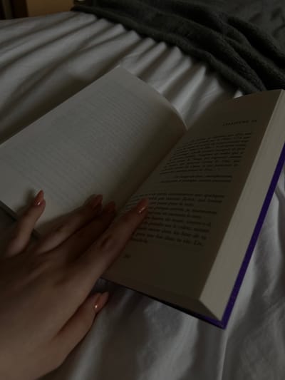 The image shows a person holding open a book with a purple cover. The text on the right page is in French, and the chapter is marked as "CHAPITRE 24". The left page is blank. The person's hand with manicured nails is visible. The background is a bed covered with white sheets and a grey blanket. There is no information to suggest the picture was taken in Truchtersheim.